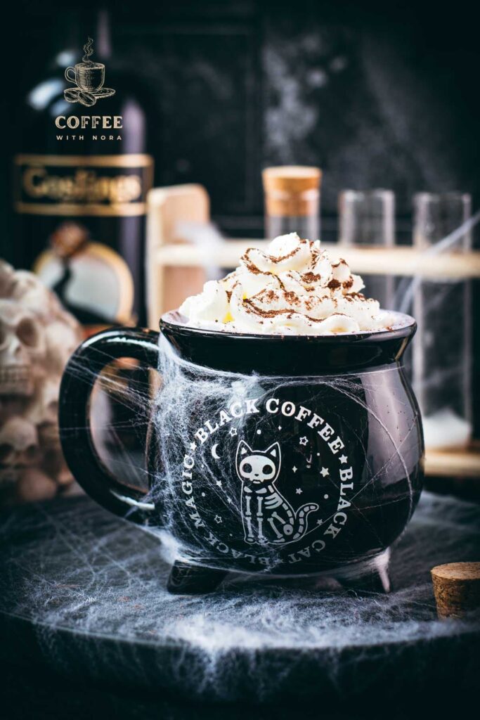 Spooky cauldron coffee mug filled with delicious coffee with rum. Topped with sweetened whipped cream and placed on a black plate surrounded by cobweb.