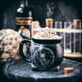 Spooky cauldron coffee mug filled with delicious coffee with rum. Topped with sweetened whipped cream and placed on a black plate surrounded by cobweb.