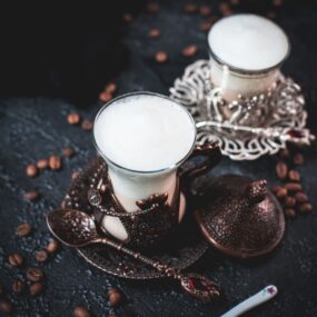 Gorgeous medieval glasses filled with delicious Breve coffee, placed on dark ground next to coffee beans.