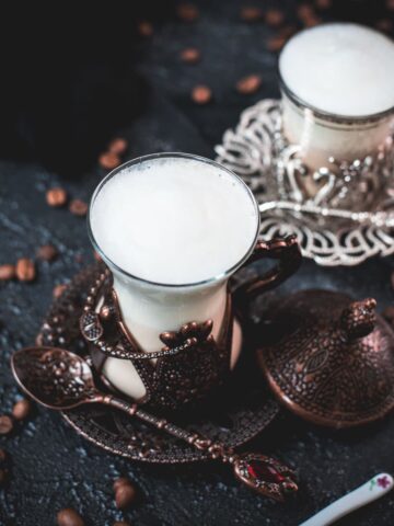 Gorgeous medieval glasses filled with delicious Breve coffee, placed on dark ground next to coffee beans.