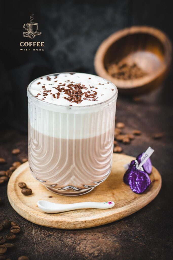 Gorgeous glass, filled with a delicious Mocha, topped with frothed milk, and garnished with chocolate sprinkles. Placed on a small wooden plate next to dark chocolate kisses.