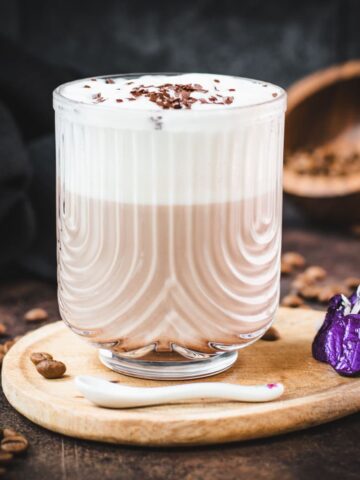 Gorgeous glass, filled with a delicious Mocha, topped with frothed milk, and garnished with chocolate sprinkles. Placed on a small wooden plate next to dark chocolate kisses.