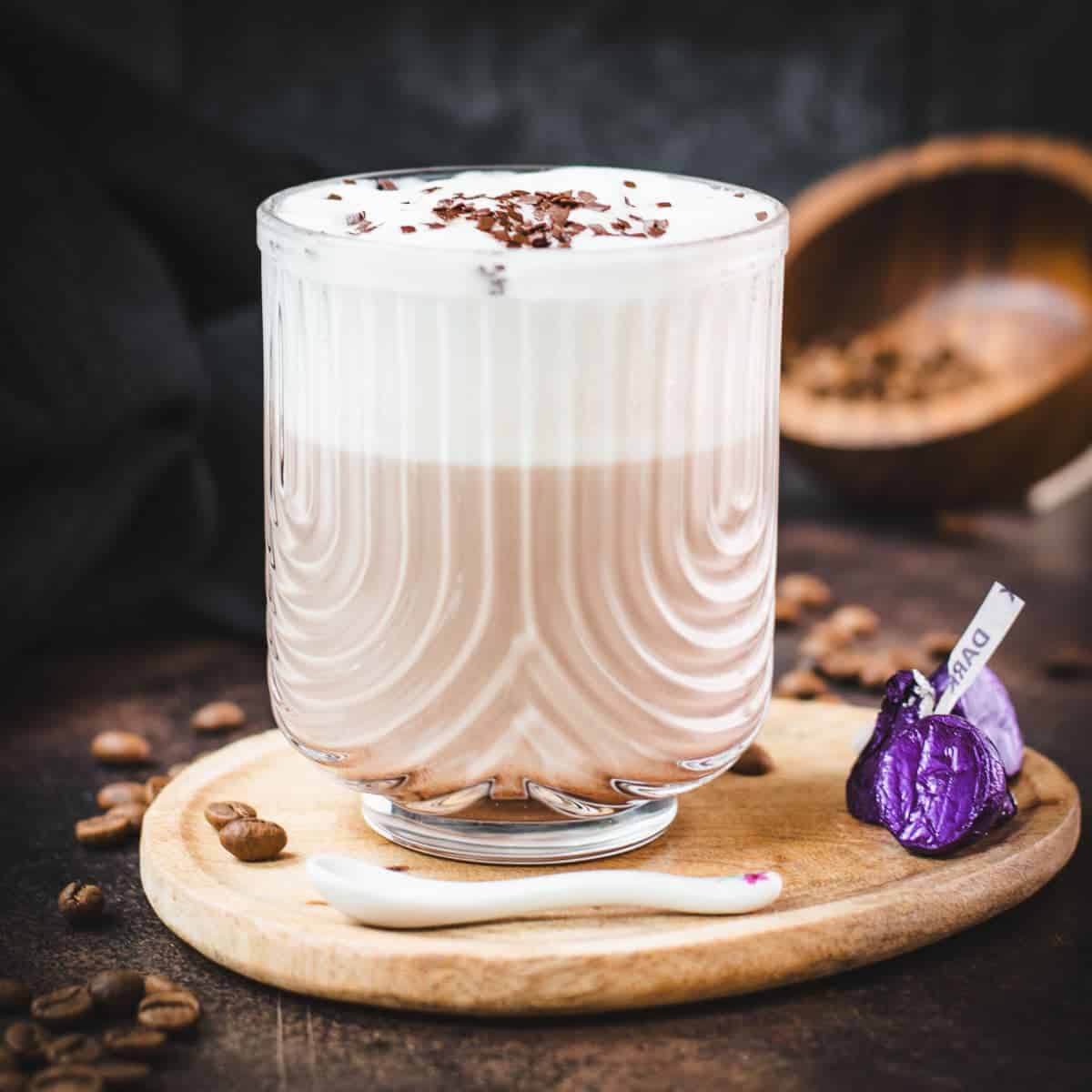 Gorgeous glass, filled with a delicious Mocha, topped with frothed milk, and garnished with chocolate sprinkles. Placed on a small wooden plate next to dark chocolate kisses.