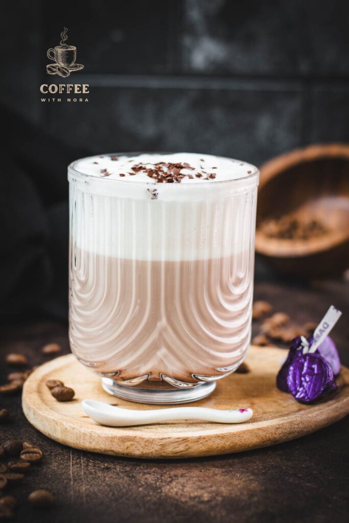 Gorgeous glass, filled with a delicious Mocha, topped with frothed milk, and garnished with chocolate sprinkles. Placed on a small wooden plate next to dark chocolate kisses.