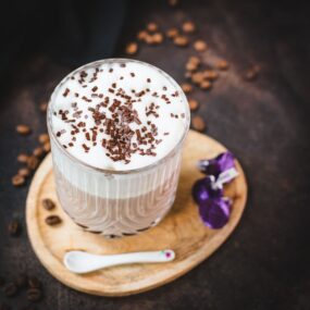 Gorgeous glass, filled with a delicious Mocha, topped with frothed milk, and garnished with chocolate sprinkles. Placed on a small wooden plate next to dark chocolate kisses.