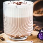 Gorgeous glass, filled with a delicious Mocha, topped with frothed milk, and garnished with chocolate sprinkles. Placed on a small wooden plate next to dark chocolate kisses.