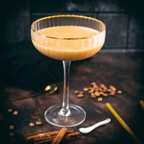 Gorgeous long stemmed cocktail glass with golden rim filled with a Cold Brew Kahlua Eggnog Cocktail, placed on dark ground.