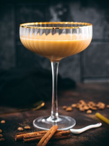 Gorgeous long stemmed cocktail glass with golden rim filled with a Cold Brew Kahlua Eggnog Cocktail, placed on dark ground.