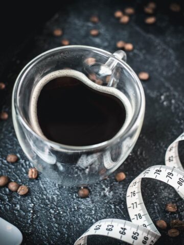 Heart-shaped coffee mug filled with black coffee, next to measuring tape on dark ground.