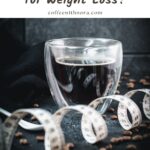 Heart-shaped coffee mug filled with black coffee, next to measuring tape on dark ground.