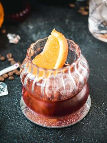 Gorgeous rocks glass, filled with a delicious cold brew negroni, garnished with an orange slice, and placed on a mirrored coaster.