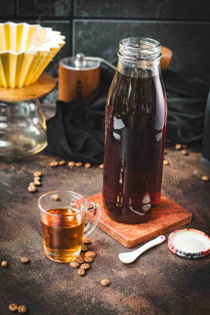 Amazing coffee syrup, in small glass next to a preserving bottle filled with coffee syrup, placed on brown ground next to coffee beans.
